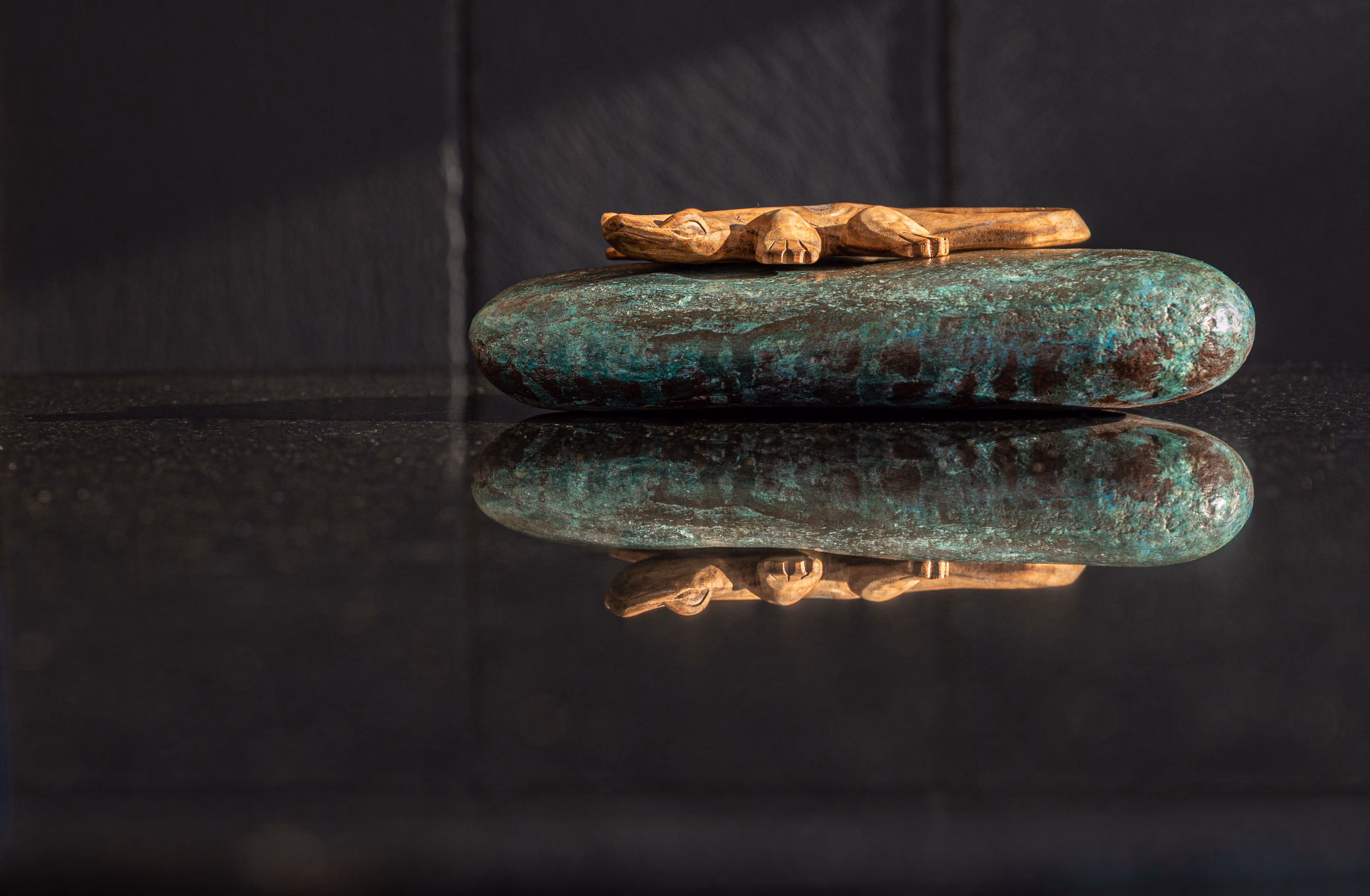 Close-up of a carved wood lizard on a patinated bronze stone, reflected in black granite.