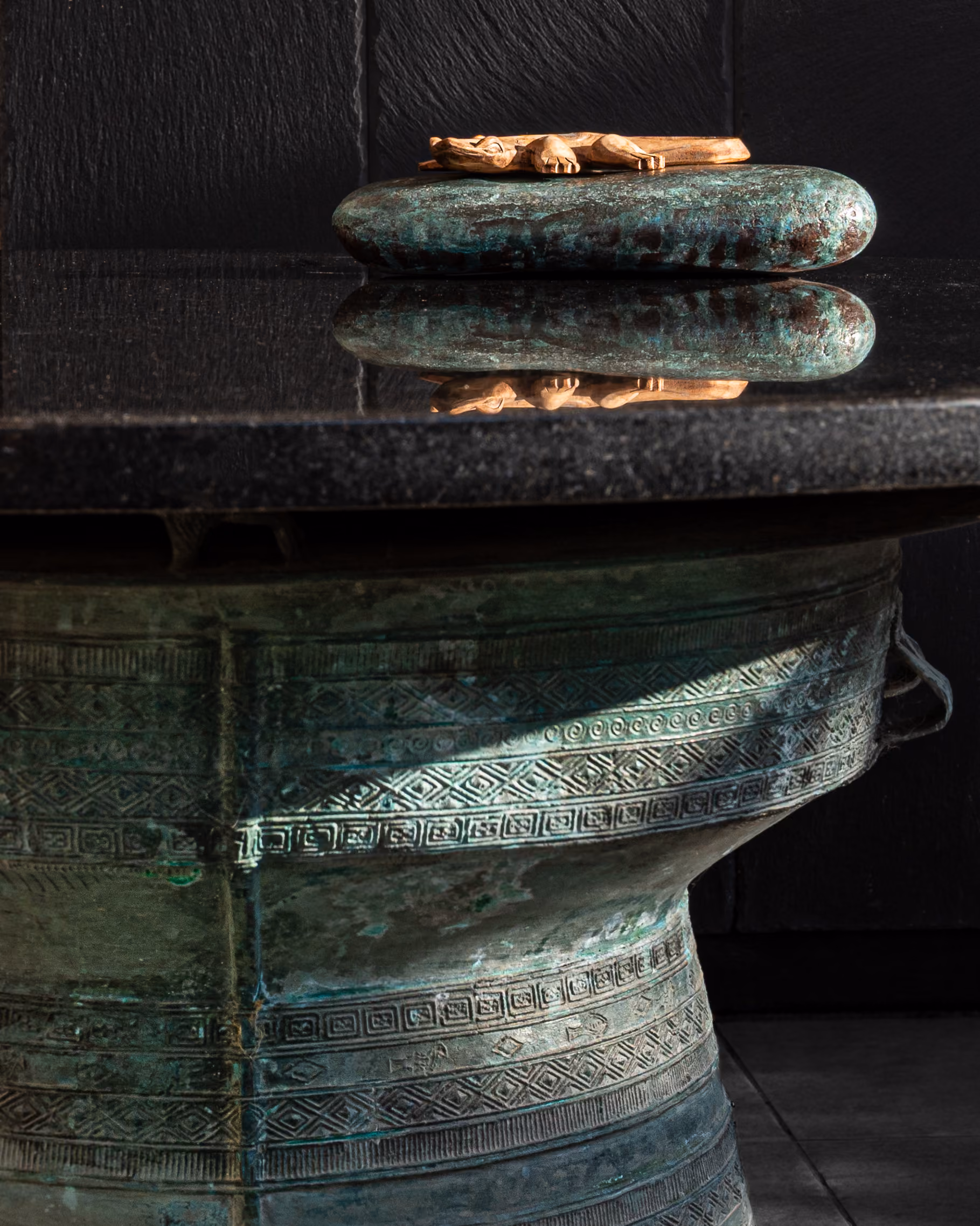 Bronze rain drum used as a table base under black granite, styled with a carved wood lizard, in a dark moody interior.